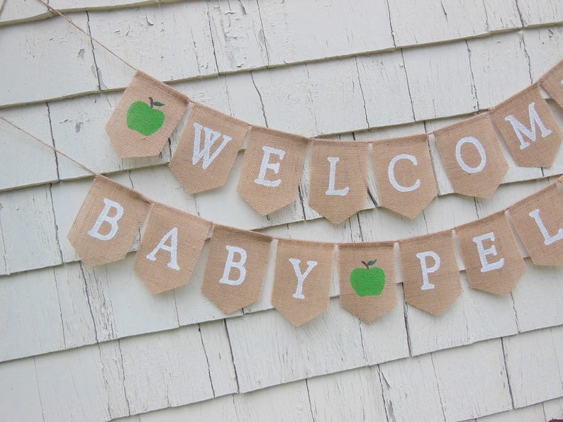K&ouml;nnte beinhalten: Ein Wimpelbanner aus Jute mit den wei&szlig;en Gro&szlig;buchstaben "WELCOME BABY". Das Banner enth&auml;lt zwei gr&uuml;ne Apfelillustrationen. Es ist an einer d&uuml;nnen Schnur befestigt und h&auml;ngt vor einem wei&szlig;en, verwitterten Holzhintergrund.