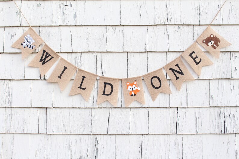 May include: A burlap banner with the words "WILD ONE" in black lettering. The banner is decorated with animal illustrations, including a raccoon, a fox, and a bear.