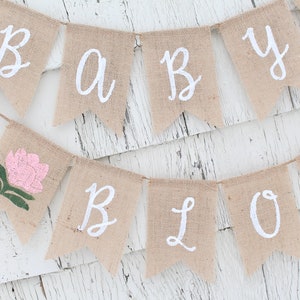 K&ouml;nnte beinhalten: Ein Banner aus Jute mit den Worten "Baby Bloom" in wei&szlig;er Farbe. Das Banner ist mit einer rosa Blume verziert.