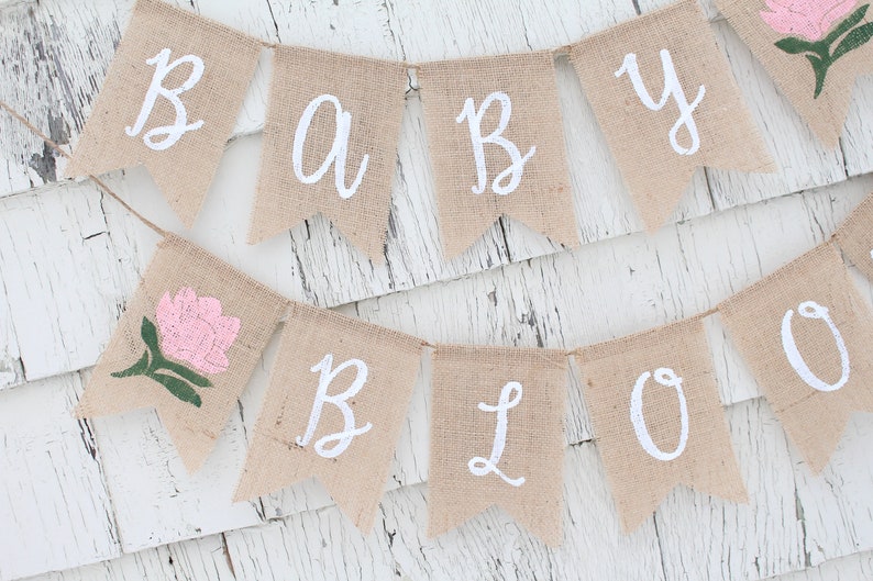 K&ouml;nnte beinhalten: Ein Banner aus Jute mit den Worten "Baby Bloom" in wei&szlig;er Farbe. Das Banner ist mit einer rosa Blume verziert.