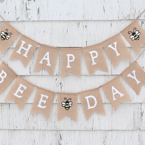 May include: A burlap banner with the words "Happy Bee Day" in white letters. The banner is decorated with three yellow and black bees.