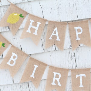 May include: A burlap banner with the words "Happy Birthday" in white paint. Each letter is on a separate flag with a small yellow lemon illustration on the top flag.