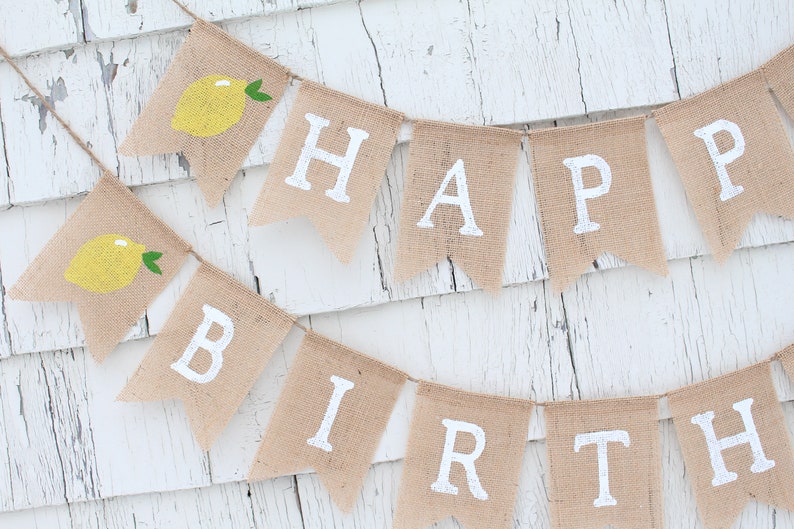 May include: A burlap banner with the words "Happy Birthday" in white paint. Each letter is on a separate flag with a small yellow lemon illustration on the top flag.