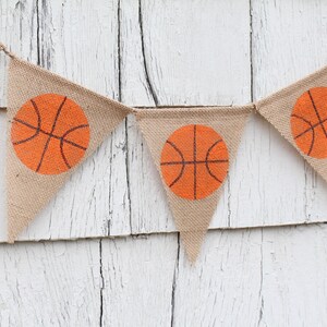 Puede incluir: Una pancarta de arpillera con balones de baloncesto naranjas impresos en cada bandera triangular. La pancarta est&aacute; colgada en una pared de madera blanca.