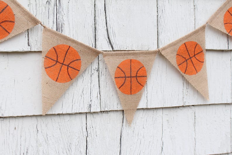 Puede incluir: Una pancarta de arpillera con balones de baloncesto naranjas impresos en cada bandera triangular. La pancarta est&aacute; colgada en una pared de madera blanca.