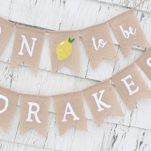 May include: A burlap banner with the words "Soon to be Drakes" in white lettering. The banner is decorated with yellow lemons and green leaves.