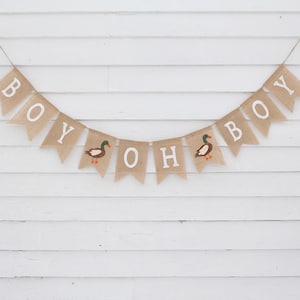May include: A burlap banner with the words "Boy Oh Boy" and two duck illustrations. The banner is strung on a white wall.