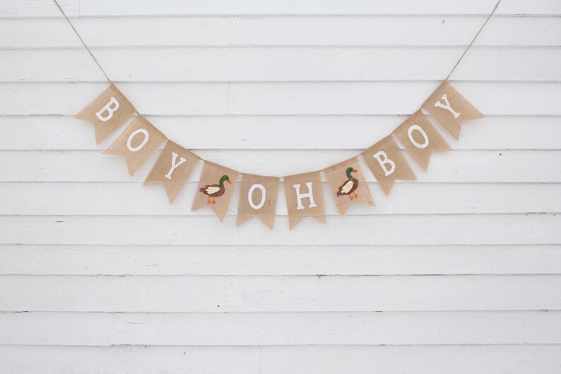 May include: A burlap banner with the words "Boy Oh Boy" and two duck illustrations. The banner is strung on a white wall.