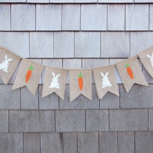 May include: A burlap banner with alternating white bunny and orange carrot silhouettes. The banner is strung on a white cord and hangs against a woodgrain background.