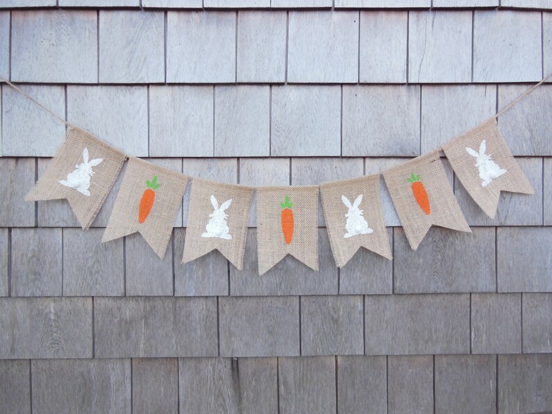May include: A burlap banner with alternating white bunny and orange carrot silhouettes. The banner is strung on a white cord and hangs against a woodgrain background.