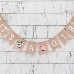 Puede incluir: Una pancarta de arpillera con las palabras "It's a Girl" en letras blancas. La pancarta est&aacute; decorada con una vaca y un cerdo.