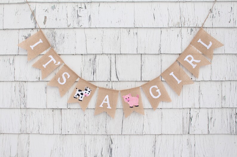 Puede incluir: Una pancarta de arpillera con las palabras "It's a Girl" en letras blancas. La pancarta est&aacute; decorada con una vaca y un cerdo.