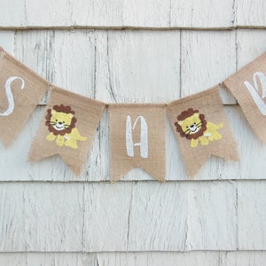 May include: A burlap banner with the words "It's a Boy" in white paint. Each letter is on a separate flag with a small lion illustration.