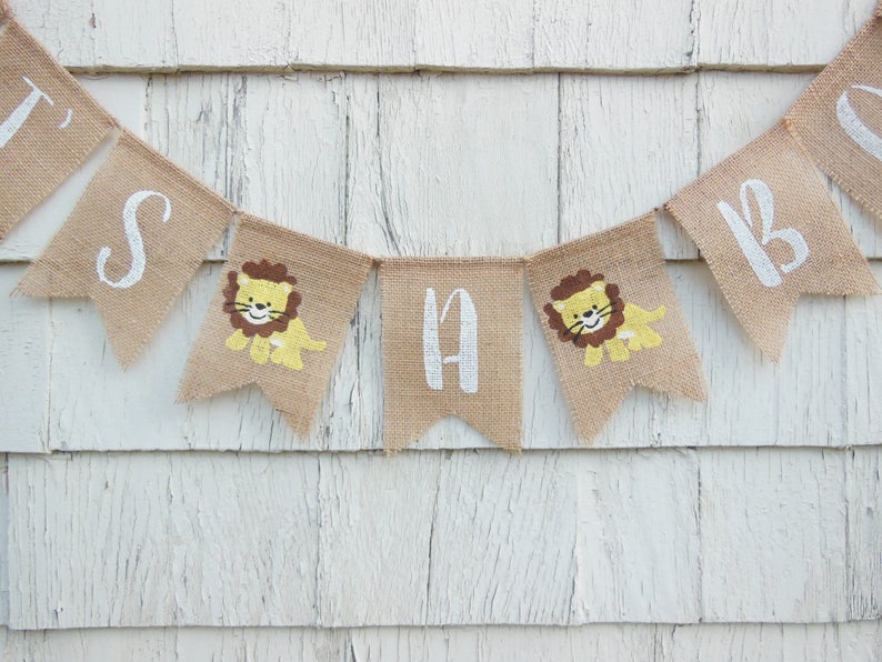 May include: A burlap banner with the words "It's a Boy" in white paint. Each letter is on a separate flag with a small lion illustration.