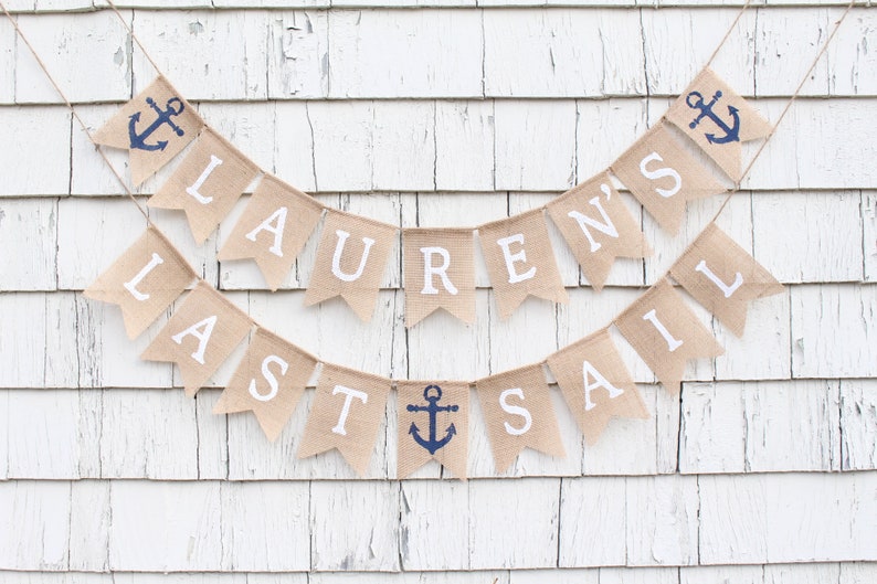 May include: A burlap banner with white letters spelling "Lauren's Last Sail" and blue anchor symbols. The banner is hanging on a white wooden wall.