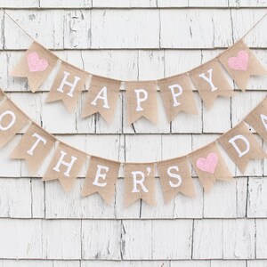 May include: A burlap banner with the words "Happy Mother's Day" in white lettering and pink hearts. The banner is strung on a white wooden wall.