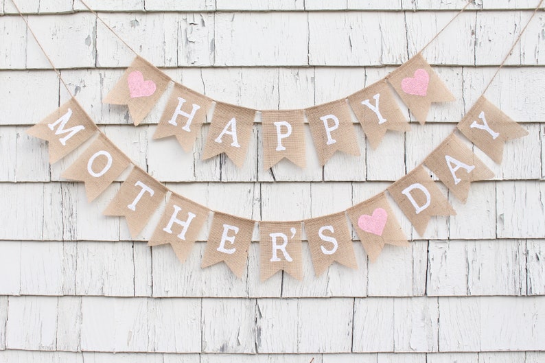 May include: A burlap banner with the words "Happy Mother's Day" in white lettering and pink hearts. The banner is strung on a white wooden wall.