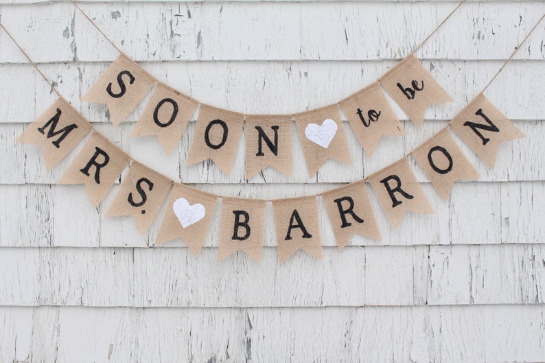 May include: A burlap banner with the text "Mrs. Soon to be Barron" in black lettering. The banner has two white hearts and is strung on a white wooden wall.