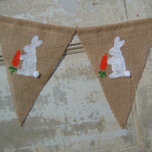 May include: A burlap banner with white bunny silhouettes holding carrots. The bunnies are outlined in white and have green leaves at their feet. The banner is strung together with white twine.