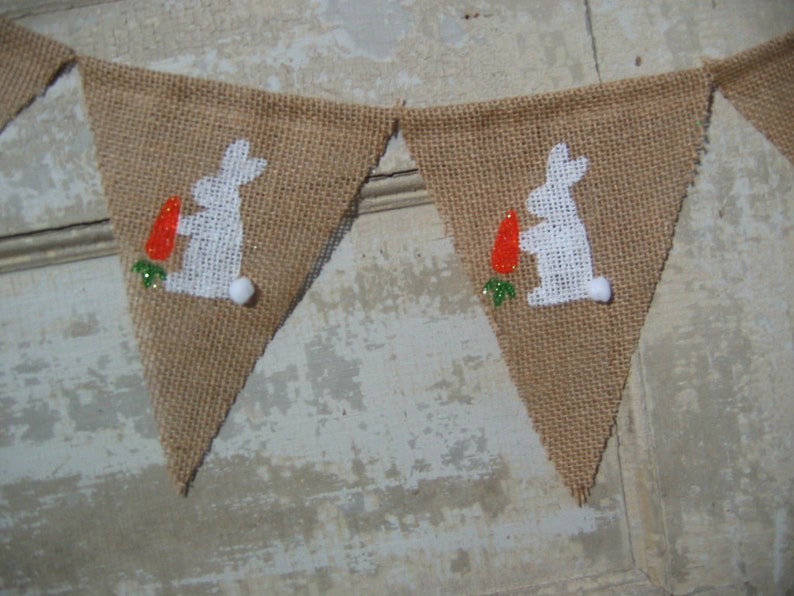 May include: A burlap banner with white bunny silhouettes holding carrots. The bunnies are outlined in white and have green leaves at their feet. The banner is strung together with white twine.