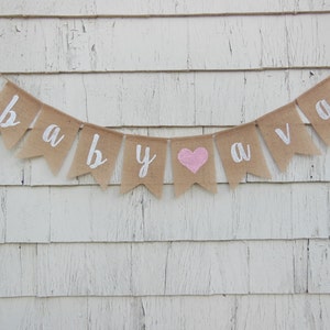 May include: A burlap banner with the words "baby ava" in white lettering. A pink heart is between the words "baby" and "ava".