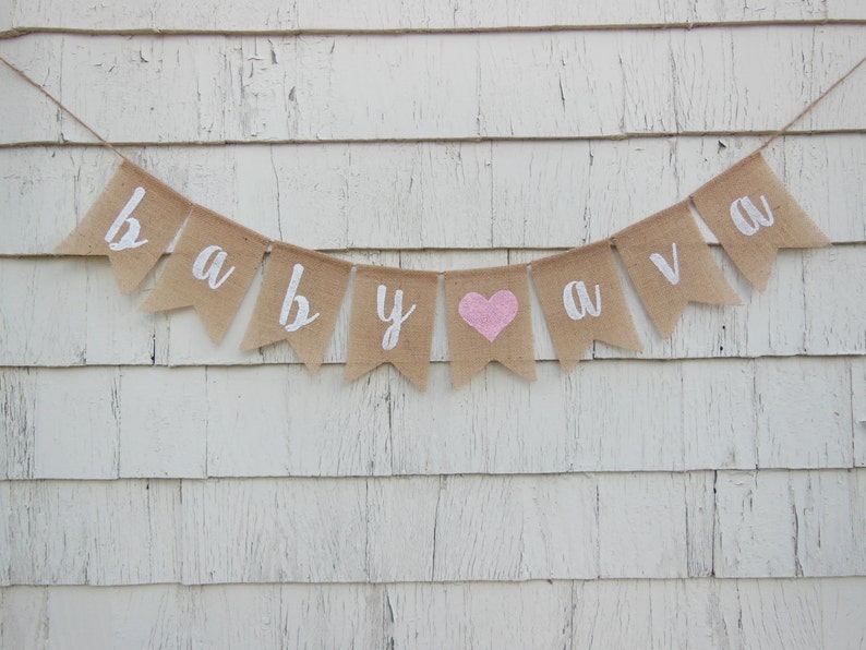 May include: A burlap banner with the words "baby ava" in white lettering. A pink heart is between the words "baby" and "ava".