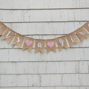 May include: A burlap banner with the text "it's a girl" in white lettering. The banner has pink hearts between the words.