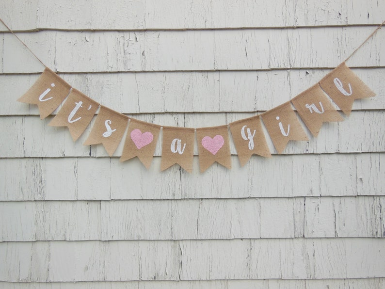 May include: A burlap banner with the text "it's a girl" in white lettering. The banner has pink hearts between the words.