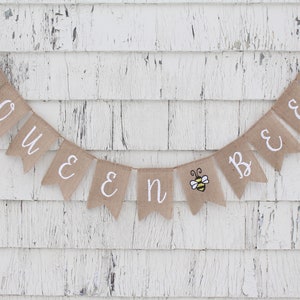 May include: A burlap banner with the words "Queen Bee" in white lettering. The banner is decorated with small yellow and black bee illustrations.