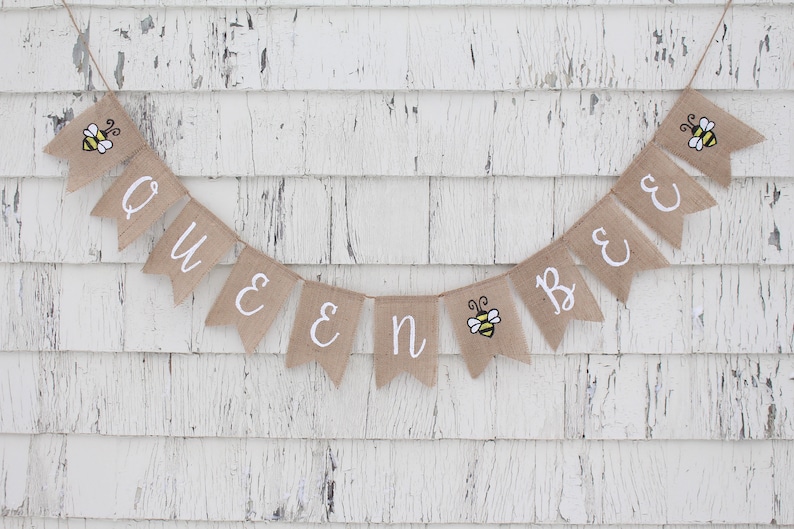 May include: A burlap banner with the words "Queen Bee" in white lettering. The banner is decorated with small yellow and black bee illustrations.