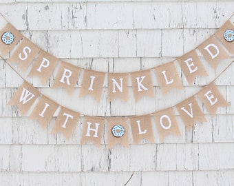 May include: A burlap banner with the text "Sprinkled with Love" in white letters. The banner is decorated with blue and brown donut images.