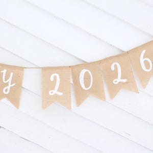 May include: A banner with pennants made of tan burlap. Each pennant has a white letter or number. The banner spells out "a y 2026". The banner is strung on a thin twine and hangs against a white wooden background.
