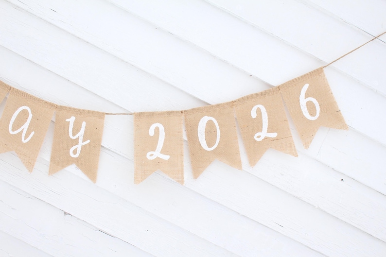 May include: A banner with pennants made of tan burlap. Each pennant has a white letter or number. The banner spells out "a y 2026". The banner is strung on a thin twine and hangs against a white wooden background.