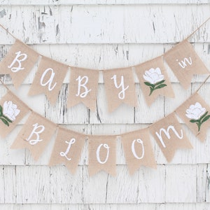 K&ouml;nnte beinhalten: Ein Banner aus Jute mit den Worten "Baby in Bloom" in wei&szlig;er Kursivschrift. Das Banner ist mit wei&szlig;en Blumen und gr&uuml;nen Bl&auml;ttern verziert.