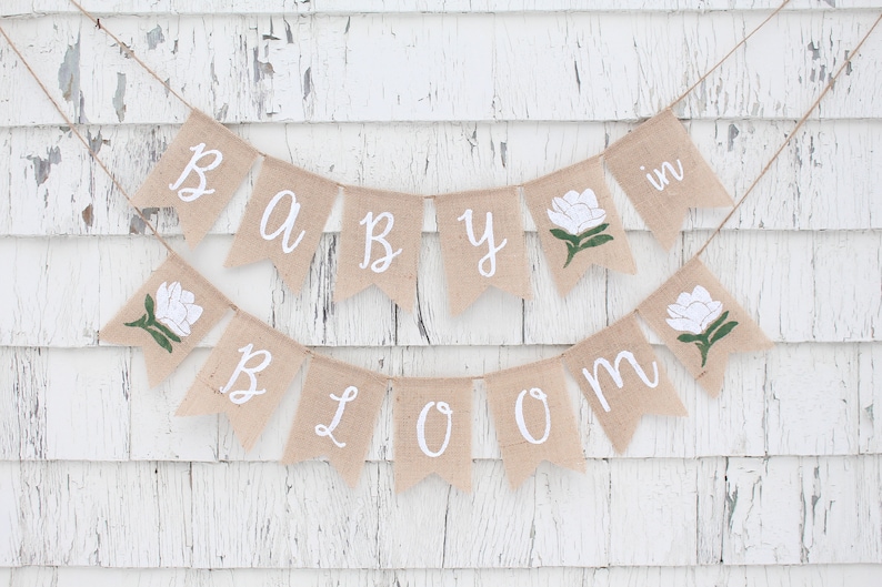 K&ouml;nnte beinhalten: Ein Banner aus Jute mit den Worten "Baby in Bloom" in wei&szlig;er Kursivschrift. Das Banner ist mit wei&szlig;en Blumen und gr&uuml;nen Bl&auml;ttern verziert.