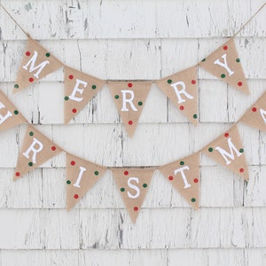 May include: A festive burlap banner spelling "MERRY CHRISTMAS" in white capital letters. Each triangular flag is decorated with red and green polka dots. The banner is suspended on twine against a white, distressed wooden backdrop.