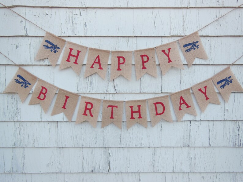 Może przedstawiać: Baner z juty z czerwonymi literami tworzącymi napis "Happy Birthday" i niebieskimi sylwetkami samolot&oacute;w. Baner jest zawieszony na białej drewnianej ścianie.