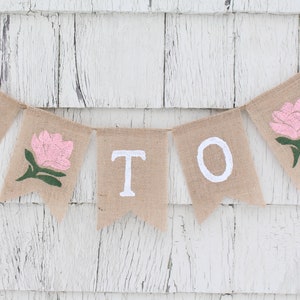 May include: A burlap banner with the word "MOM" spelled out in white paint. Each letter is on a separate flag, and there are pink flowers with green stems between the letters.