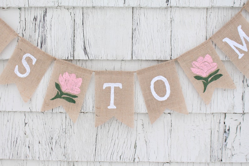 May include: A burlap banner with the word "MOM" spelled out in white paint. Each letter is on a separate flag, and there are pink flowers with green stems between the letters.