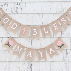 Puede incluir: Una pancarta de arpillera con las palabras "God Bless Maya" en letras blancas. La pancarta est&aacute; decorada con flores rosas y una cruz blanca.