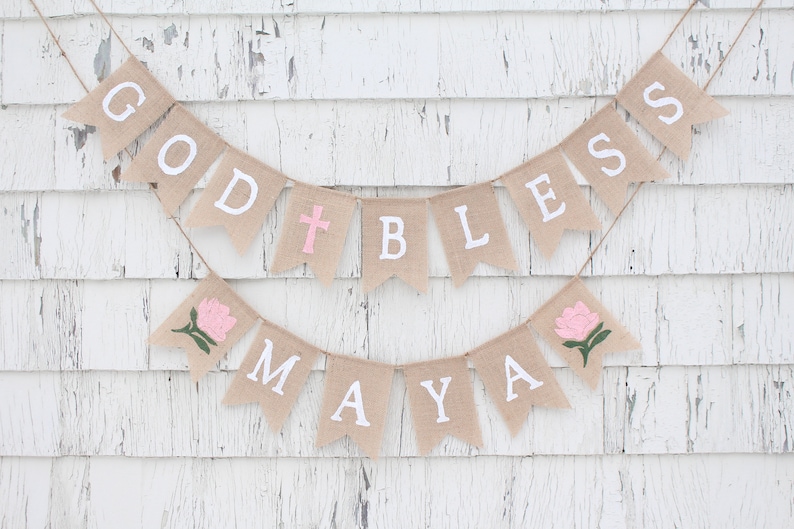 Puede incluir: Una pancarta de arpillera con las palabras "God Bless Maya" en letras blancas. La pancarta est&aacute; decorada con flores rosas y una cruz blanca.