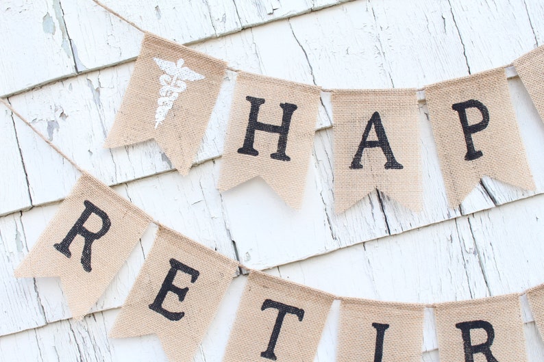May include: A burlap banner with the words "Happy Retirement" in black lettering. The banner is strung on a white wooden wall. A small burlap flag with a white medical symbol is at the beginning of the banner.