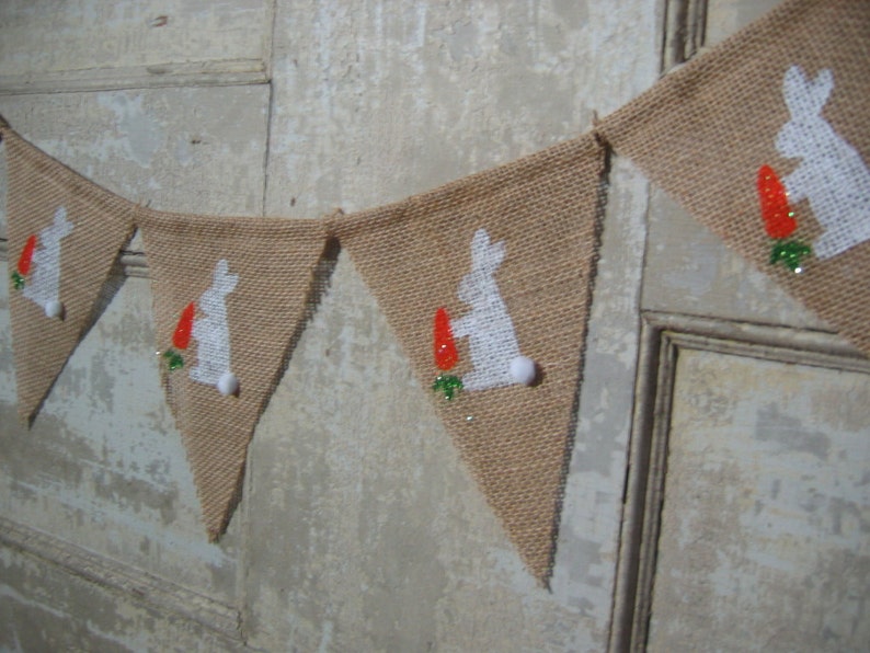 May include: A burlap banner with white bunny silhouettes and orange carrots. The banner is strung together with white pom-poms.