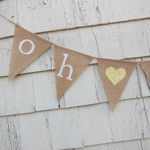 May include: A burlap banner with the words "Oh Baby" in white letters and a yellow heart in the center. The banner is hanging on a white wooden wall.