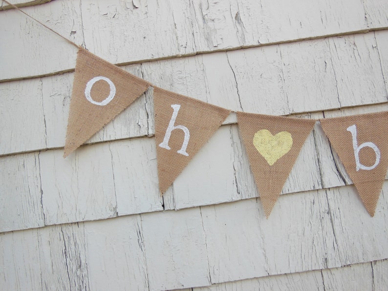 May include: A burlap banner with the words "Oh Baby" in white letters and a yellow heart in the center. The banner is hanging on a white wooden wall.