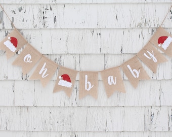 May include: A burlap banner with the words "oh baby" in white lettering. Each pennant has a red Santa hat with white trim. The banner is strung on twine and is against a white, weathered wood background.