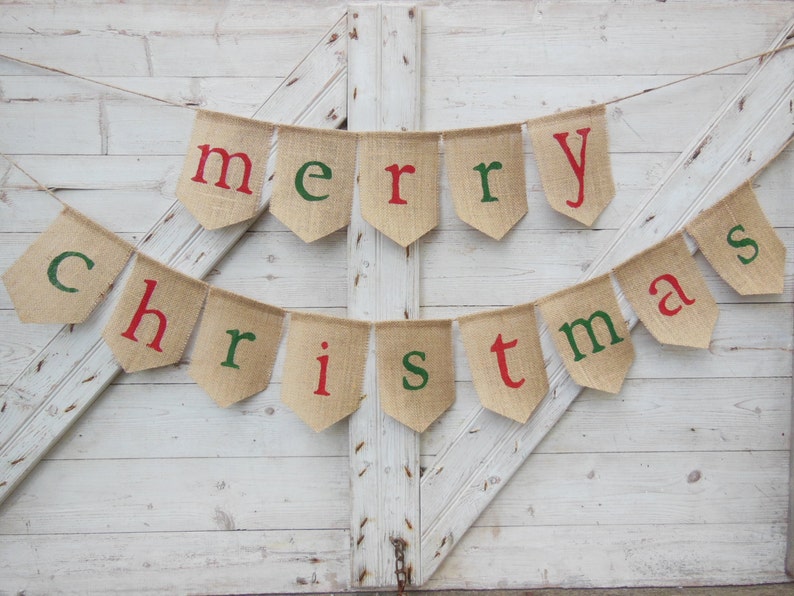 K&ouml;nnte beinhalten: Ein Banner aus Jute mit den Worten "merry christmas" in roten und gr&uuml;nen Buchstaben. Der Banner h&auml;ngt vor einem wei&szlig;en Holzhintergrund und ist eine rustikale Weihnachtsdekoration.