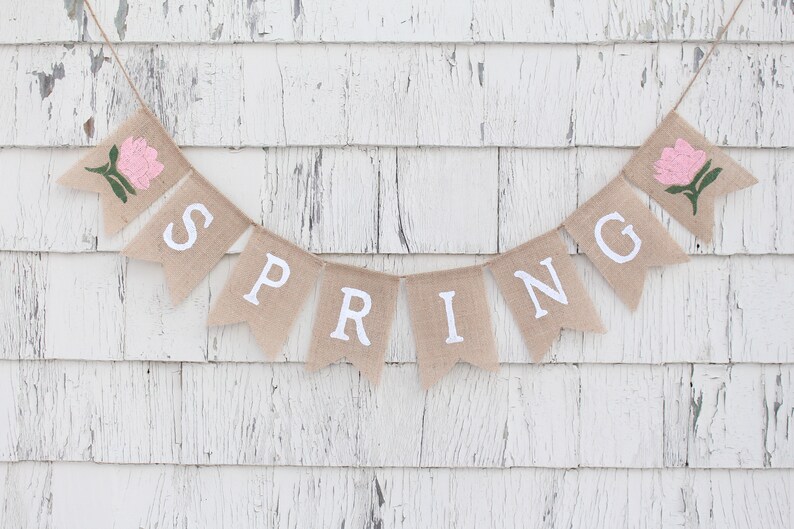 May include: A burlap banner with the word "SPRING" spelled out in white letters. The banner is decorated with two pink flowers.