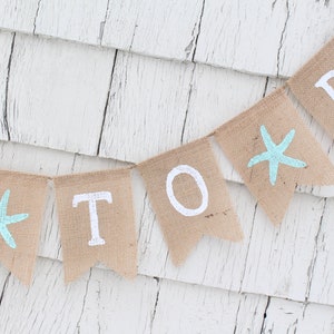 May include: A burlap banner with the words "To Be" in white paint. The banner is decorated with blue starfish.