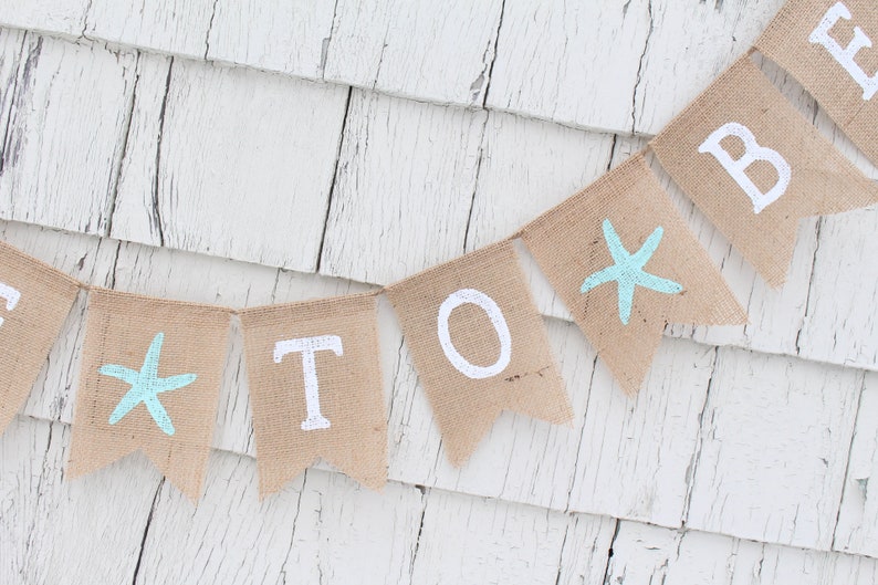 May include: A burlap banner with the words "To Be" in white paint. The banner is decorated with blue starfish.
